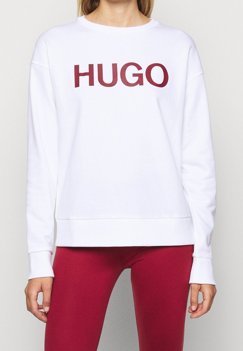 Vit sweatshirt med en framträdande vinröd "HUGO"-logotyp. Den har en avslappnad passform, långa ärmar och ribbad nederkant och muddar.