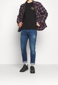 Rutig flanellskjorta i lila och grått över en svart t-shirt. Blå jeans med uppvikta ärmar och svarta sneakers fulländar outfiten.