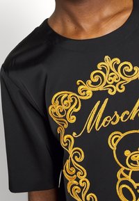 Musta puuvillatee, jossa on koristeellinen kultainen brodeeraus, mukaan lukien kiehtova kuvio ja nimi "Moschino". Lyhyet hihat ja pyöreä kaula-aukko.