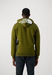 Olijfgroene hoodie met een voorste kangoeroezak, ribbels aan de mouwen en onderkant. Beschikt over een groot wit logo op de capuchon. Zachte stoftextuur.