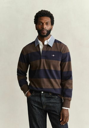 Homme barbu portant un maillot de rugby rayé marron et bleu marine par-dessus une chemise blanche à col et un jean foncé, debout avec une main dans la poche.