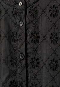 Schwarzes besticktes Hemd mit Knopfverschlüssen, das florale Muster und einen strukturierten Stoff aufweist. Die Muster bilden ein durchgehendes Diamantdesign.