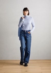 Light blue turtleneck sweater, high-waisted blue jeans, and black shoes. Sweater draped over left arm; minimal accessories.