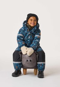 Lapsi istuu pehmeän harmaan sian päällä, pukeutuneena siniseen, eläinkuvioiseen vedenpitävään takkiin, keltaisiin lapasiin ja mustiin kenkiin.