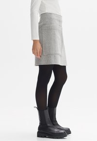 Personne portant une jupe grise avec une grande poche devant, des collants noirs, des bottes noires épaisses et un haut blanc à manches longues, debout de côté.