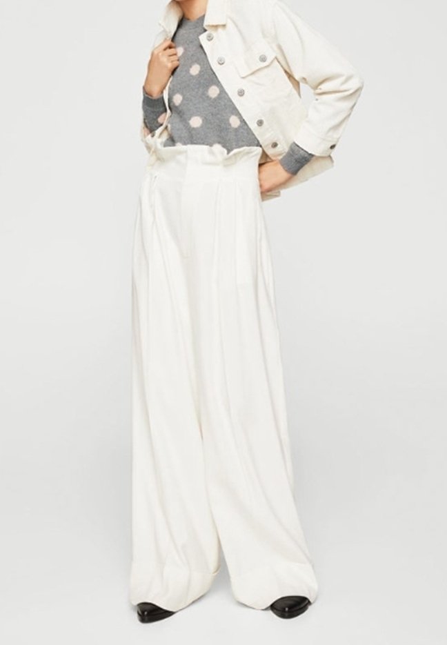 Personne portant un pantalon blanc à jambes larges, un pull gris à pois blancs, une veste courte blanche et des chaussures noires.