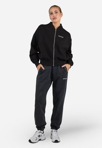 Felpa nera con zip, maniche e vita a coste, abbinata a pantaloni della tuta grigi scuri con logo. Sneakers bianche completano il look.