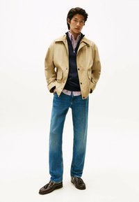 Giacca leggera beige, maglione blu navy, camicia a righe, jeans blu e mocassini scuri. Texture morbide con vestibilità casual e look a strati.