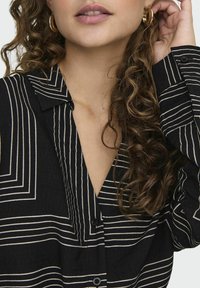 Zwarte shirt met witte verticale strepen, kraagontwerp, knoopsluiting en textuurstof. Model heeft krullend haar en gouden ringoorbellen.