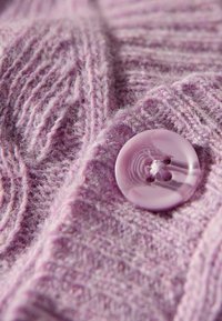 Lavendelfarbene Strickjacke mit strukturierten Zopfmustern, versehen mit einem runden, glänzenden lila Knopf mit vier Löchern, im Nahaufnahme präsentiert.