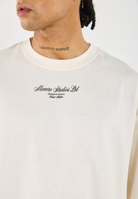 Cream-colored t-shirt featuring a round neckline and embroidered logo in black text, with a soft, smooth texture.