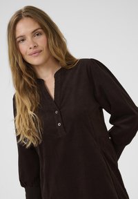 Bruine corduroy tuniek met een knoopsluiting bij de henley-kraag, lange pofmouwen en zijzakken. Zachte textuur met zichtbare geribbelde structuur.