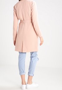 Trench coat rosa chiaro con vita regolabile, maniche lunghe e apertura posteriore. Abbinato a jeans azzurri arrotolati e sneakers bianche.