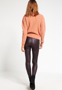 Žena s vlasy po ramena, oblečená v brokátové svetru, tmavohnědých kožených kalhotách a černých kotníkových botách, stojící na pozadí jedné bílé stěny.