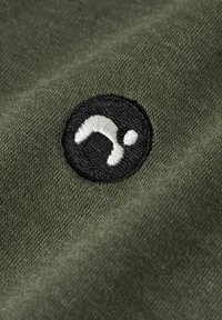 Zielony materiał o gładkiej teksturze ma czarne okrągłe haftowane logo, przedstawiające białą abstrakcyjną grafikę.