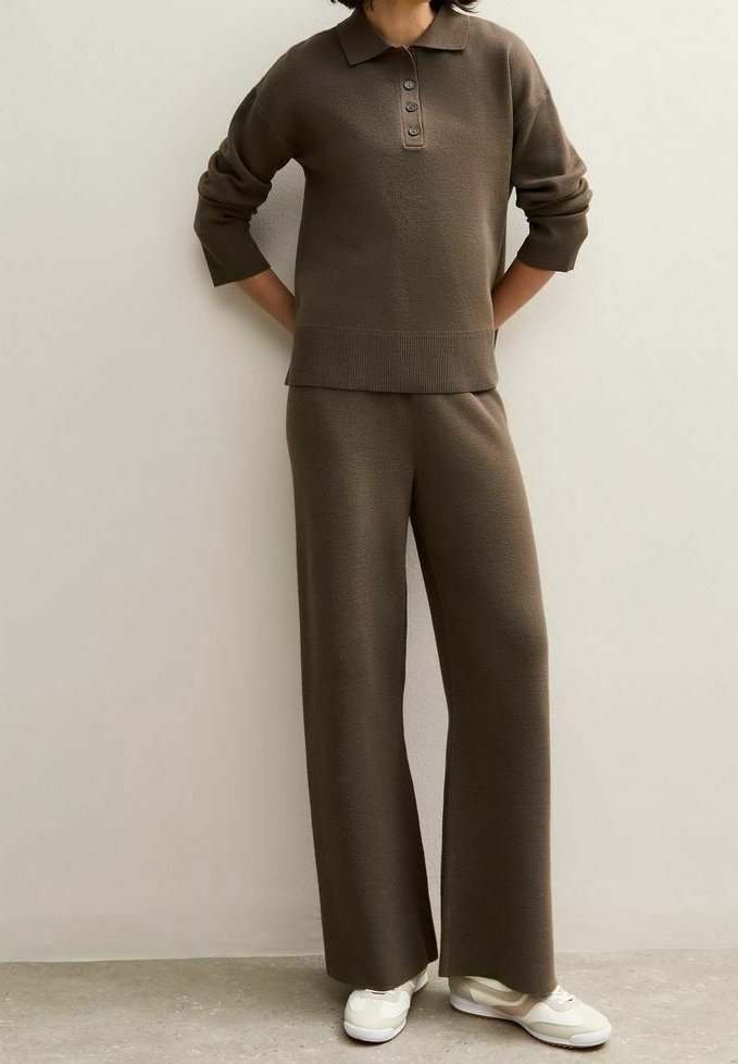 Personne portant un polo en maille marron à manches longues et un pantalon en maille large assorti avec des baskets blanches, debout contre un mur uni.
