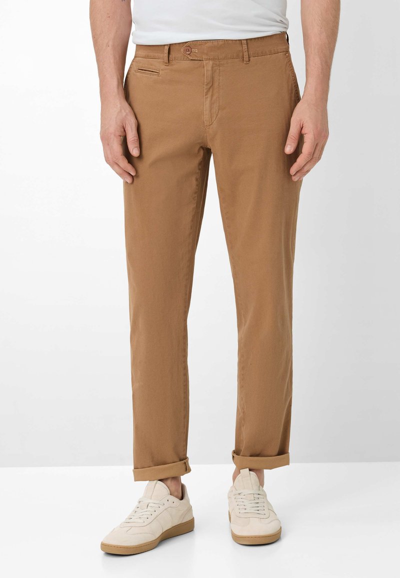 Mann trägt hochgekrempelte braune Hose und weiße Sneaker, steht vor einem schlichten hellen Hintergrund.