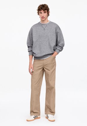 Jeune homme debout, portant un sweat gris ample, un pantalon beige à jambes larges et des baskets blanches avec des bandes noires sur un fond uni.