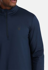 Marineblaues Langarmshirt mit hohem Kragen und halber Reißverschlussgestaltung; verfügt über ein dezentes Spinnensymbol in dunklem Farbton auf der Brust. Glatte Stofftextur.