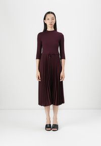 Mauve midi-kjole med ribbet body, tre kvart ermer, plissert skjørt, og snøring i midjen. Brukt med svarte sandaler.
