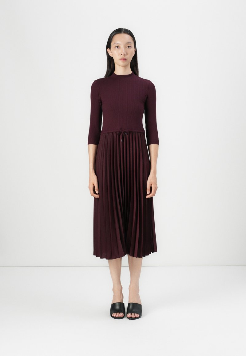 Mauve midi-kjole med ribbet body, tre kvart ermer, plissert skjørt, og snøring i midjen. Brukt med svarte sandaler.
