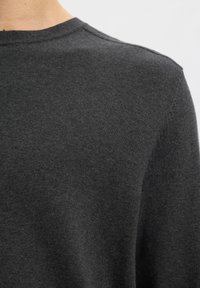 Primer plano de una persona que lleva un jersey de punto gris oscuro, centrado en el hombro y el escote con un fondo liso.