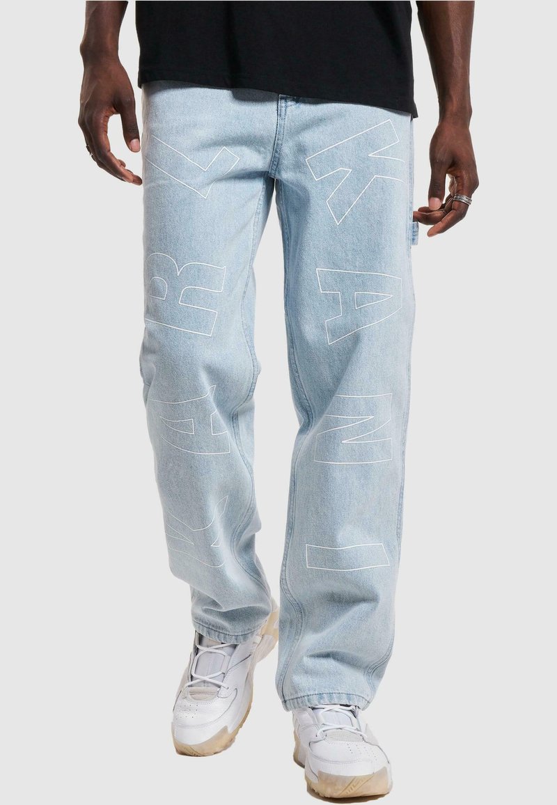 Lyseblå denimbukser med store hvite blokkbokstaver over bena. Designet med en avslappet passform og sydd detaljer, kombinert med hvite joggesko.