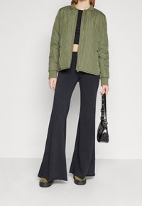 Quiltad olivgrön bomberjacka med ribbade manschetter, i kombination med svarta utsvängda byxor och en svart handväska, har en minimalistisk design.