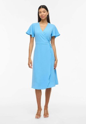 Femme portant une robe portefeuille bleu clair à manches papillon et des sandales à talons hauts transparentes, debout devant un fond blanc.