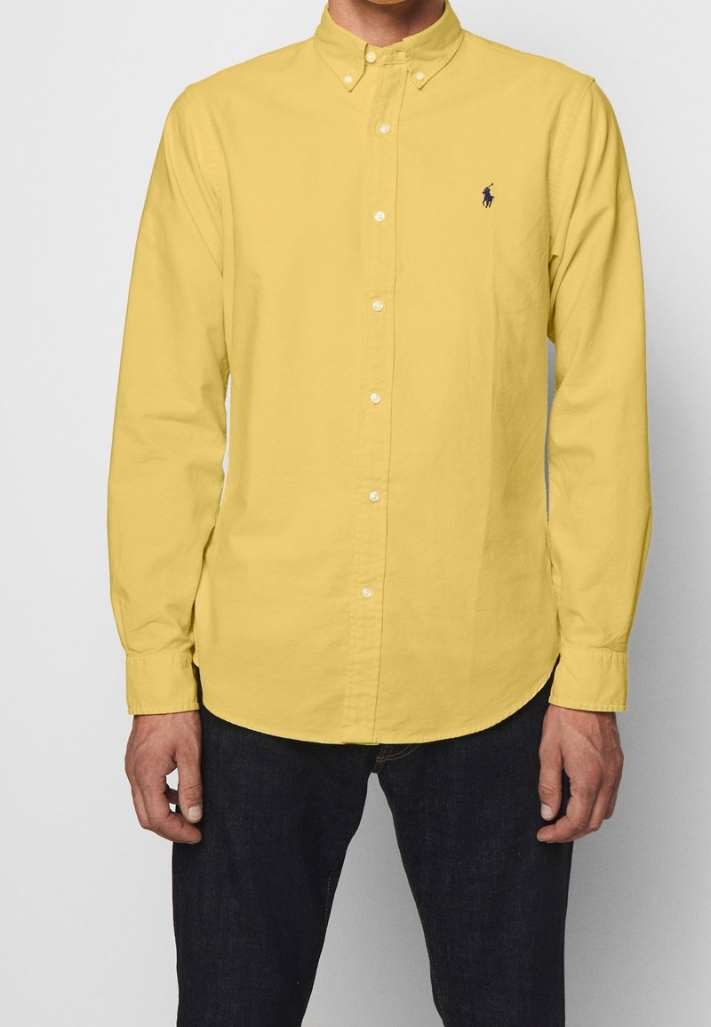 Man som bär en gul långärmad skjorta med knappar och en liten broderad mörkblå polo-spelarlogotyp på bröstet, tillsammans med mörka jeans.