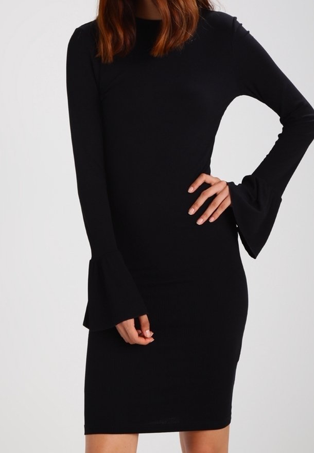 Femme portant une robe noire près du corps avec de longues manches cloche, posant avec une main sur la hanche et l'autre reposant vers le bas.