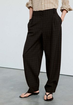 Persona que lleva pantalones oscuros a cuadros sueltos con las manos en los bolsillos, camisa a cuadros beige con las mangas remangadas y sandalias negras tipo chancla.