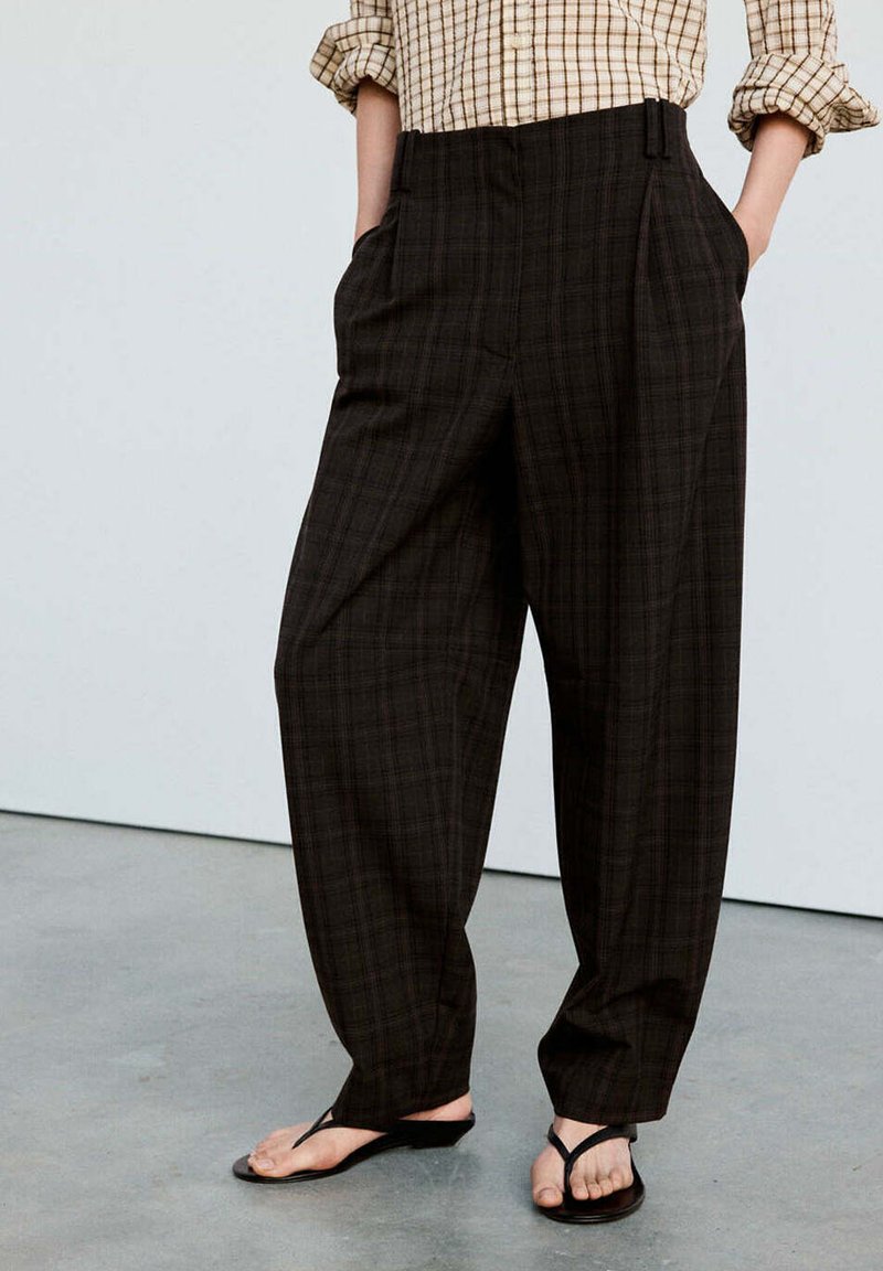 Personne portant un pantalon ample à carreaux foncés avec les mains dans les poches, une chemise à carreaux beige aux manches retroussées et des sandales noires à lanière entre les orteils.