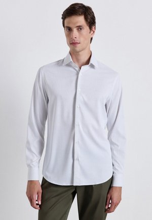 Giovane uomo che indossa una camicia bianca a maniche lunghe abbottonata e pantaloni verde oliva, in piedi davanti a uno sfondo chiaro e semplice.