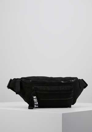 Sac banane en tissu noir avec des sangles réglables et deux poches avant zippées, une tirette de fermeture éclair étiquetée "TURN,", exposée sur un socle blanc.