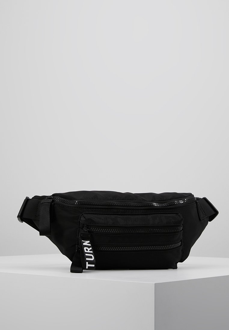 Sac banane en tissu noir avec des sangles réglables et deux poches avant zippées, une tirette de fermeture éclair étiquetée "TURN,", exposée sur un socle blanc.