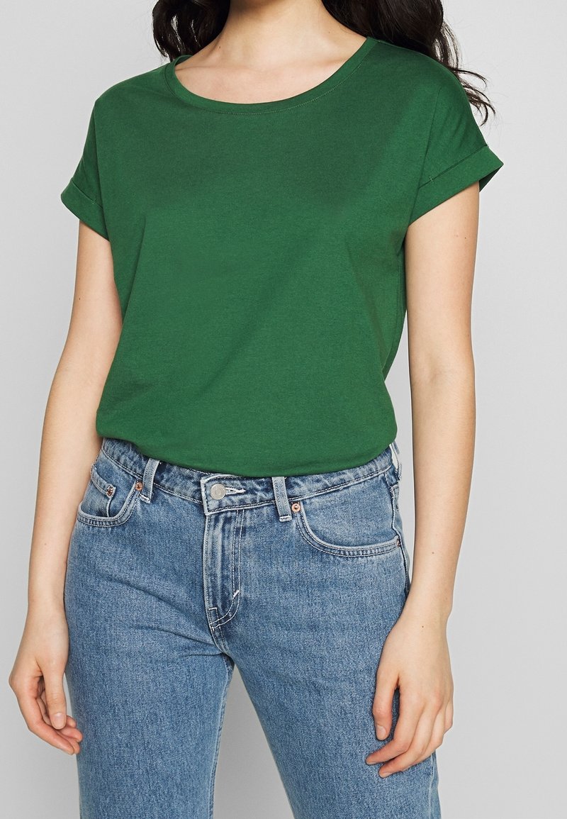 Femme portant un T-shirt vert uni à manches courtes rentré dans un jean bleu clair taille haute, sur un fond neutre.