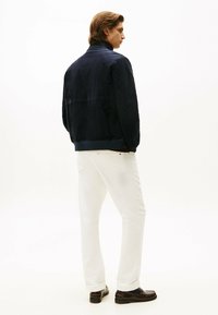 Cazadora bomber de ante azul marino con puños acanalados, combinada con jeans de mezclilla blancos y mocasines marrón oscuro, vista desde atrás.