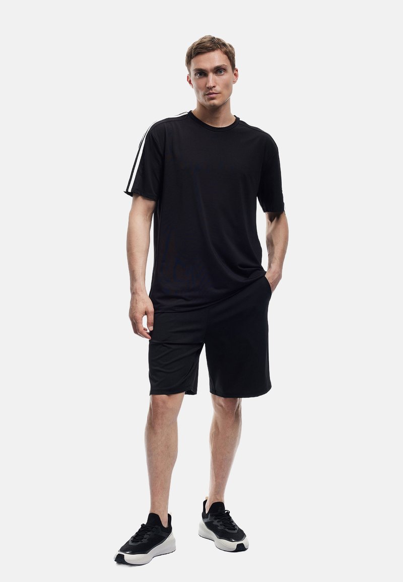 Schwarzes Sport-T-Shirt mit kurzen Ärmeln und weißen Seitenstreifen, kombiniert mit schwarzen Shorts. Leichtes Material, klassischer Rundhalsausschnitt und figurbetonter Schnitt.