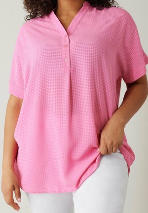 Mujer con una blusa rosa texturizada de manga corta y suelta con tres botones sobre pantalones blancos, mano sujetando el dobladillo de la blusa.