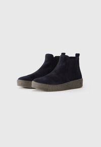 Une paire de bottes noires en daim à la cheville avec des semelles en caoutchouc épais et crantées, et des panneaux élastiques sur les côtés, sur fond blanc.