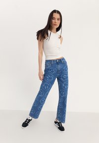 Jeans de denim azul con un gran estampado de cara sonriente, de cintura alta y corte recto. Combinados con una camiseta sin mangas blanca ajustada y zapatillas negras.