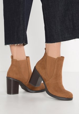 Brune ruskind ankle støvler med blokhæle, båret med mørke, korte jeans mod en helt hvid baggrund.