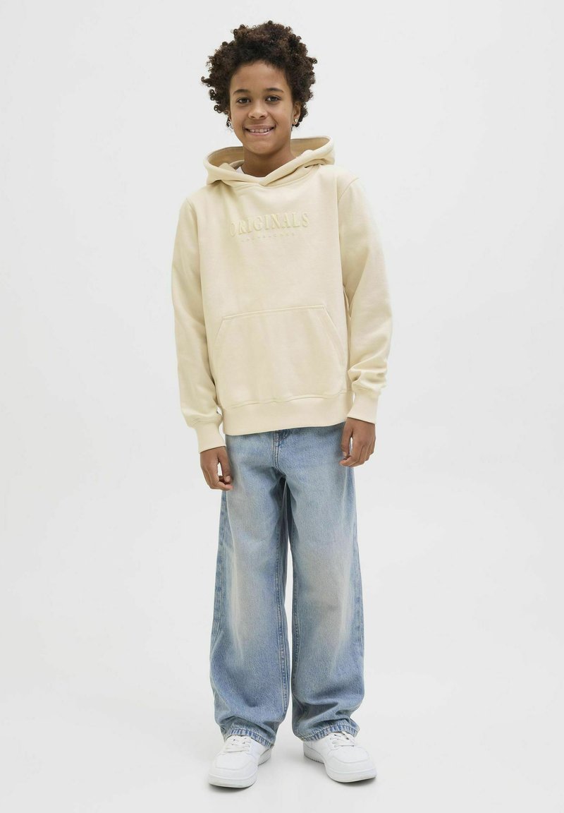 Bežs kapučja krekers ar priekšējo kabatu un izspiežamu "Originals" tekstu, apvienots ar gaiši ziliem platām džinsām un baltām kedām. Vienkāršs fons.