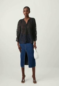 Nais, kes kannab musta pitsbluusit, sinist põlve pikkust denim-seelikut esi-lõhega, pruune sandaale ja hoiab valget käekotti.