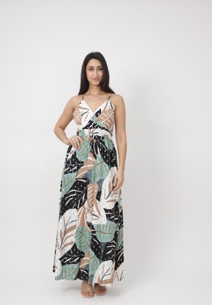 Femme en robe longue multicolore sans manches et à col en V avec un motif de feuilles, debout avec une main sur la hanche devant un fond blanc uni.