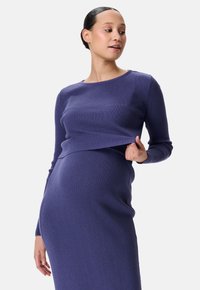 Zwangere vrouw die haar geribbelde paarse trui oplicht om haar babybuik te onthullen, met een opgestoken haar en een neutrale achtergrond.