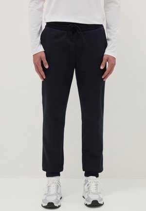 Hombre vistiendo pantalones joggers negros con puños elásticos, camisa blanca de manga larga, calcetines blancos y zapatillas deportivas blancas, de pie frente a un fondo liso.