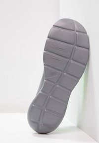 Sole of grey Nike shoe resting against white wall, showing textured tread pattern and Nike logo in center.