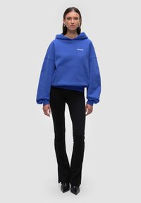 Felpa oversize blu con tasca frontale e orlo elastico, abbinata a pantaloni neri a campana e tacchi neri a punta. Tessuto morbido.